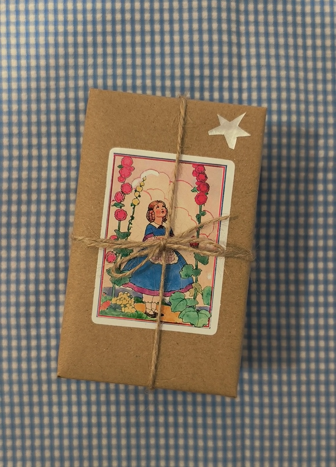 A small kraft paper gift box tied with twine and featuring a vintage-style illustration sticker, placed on a blue and white checkered fabric background.