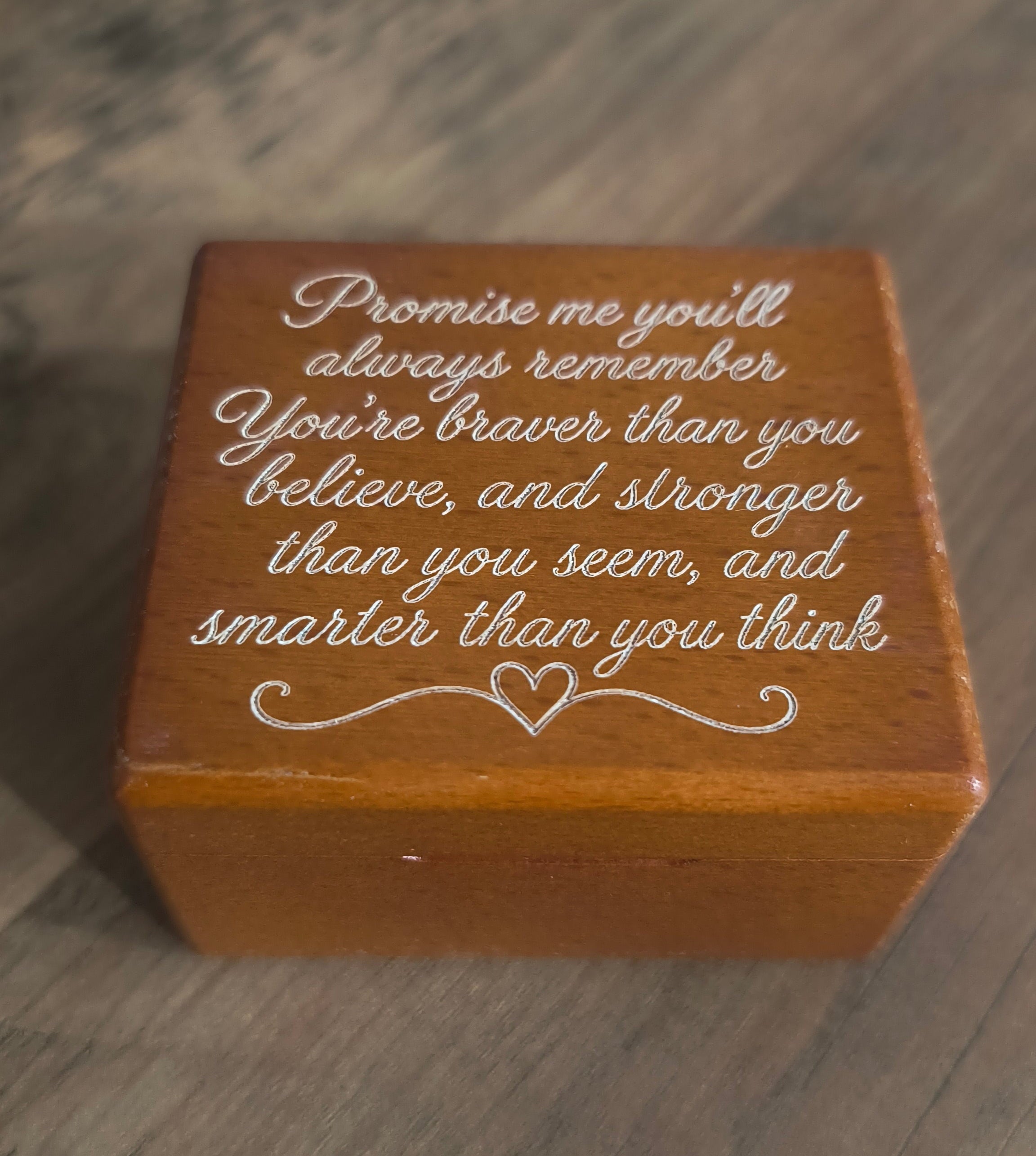 A square wooden keepsake box with engraved white script text, resting on a gray wood-textured surface.