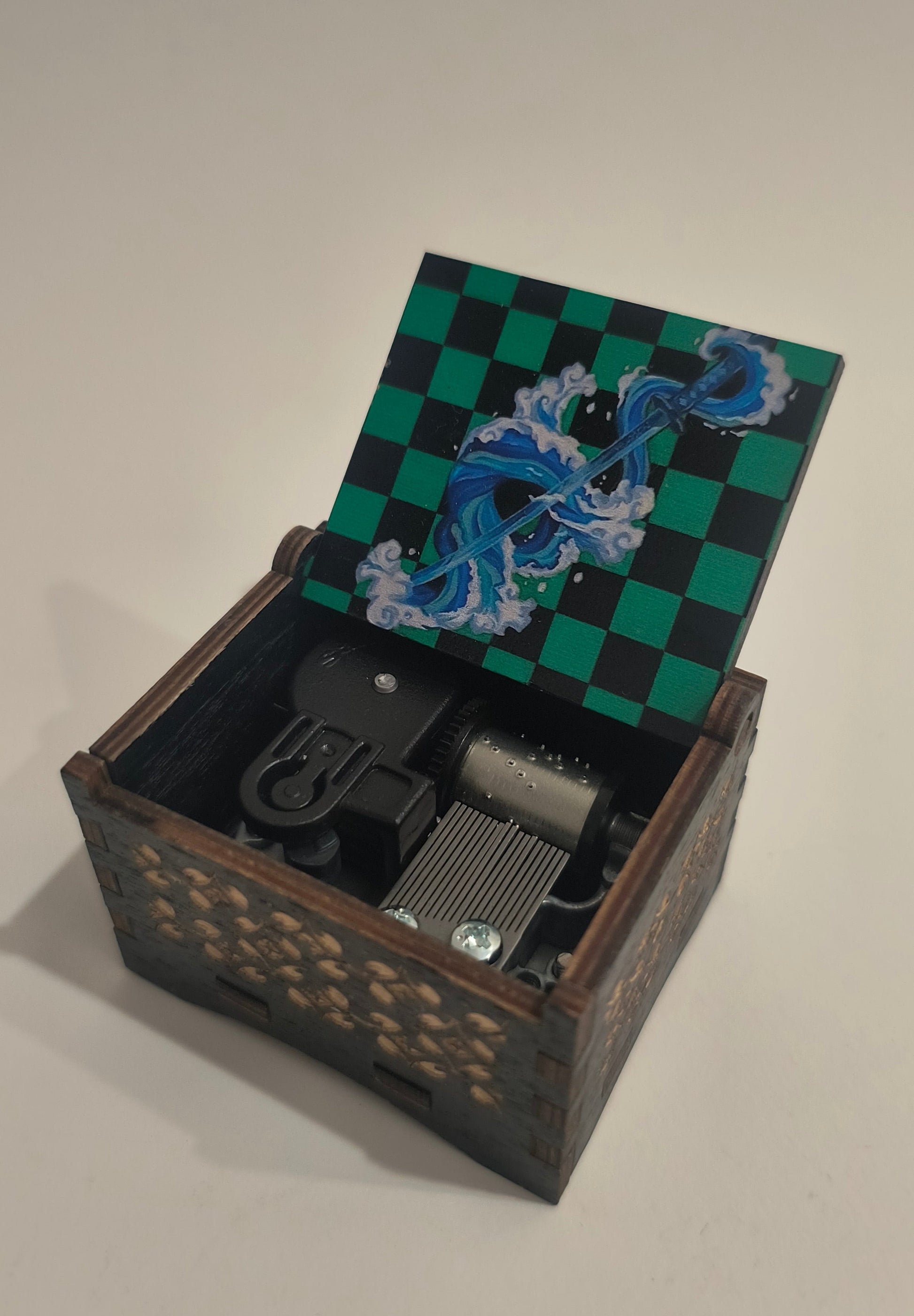 A wooden music box with a decorative green and black checkered lid featuring blue and white wave illustrations, shown open against a plain light background.