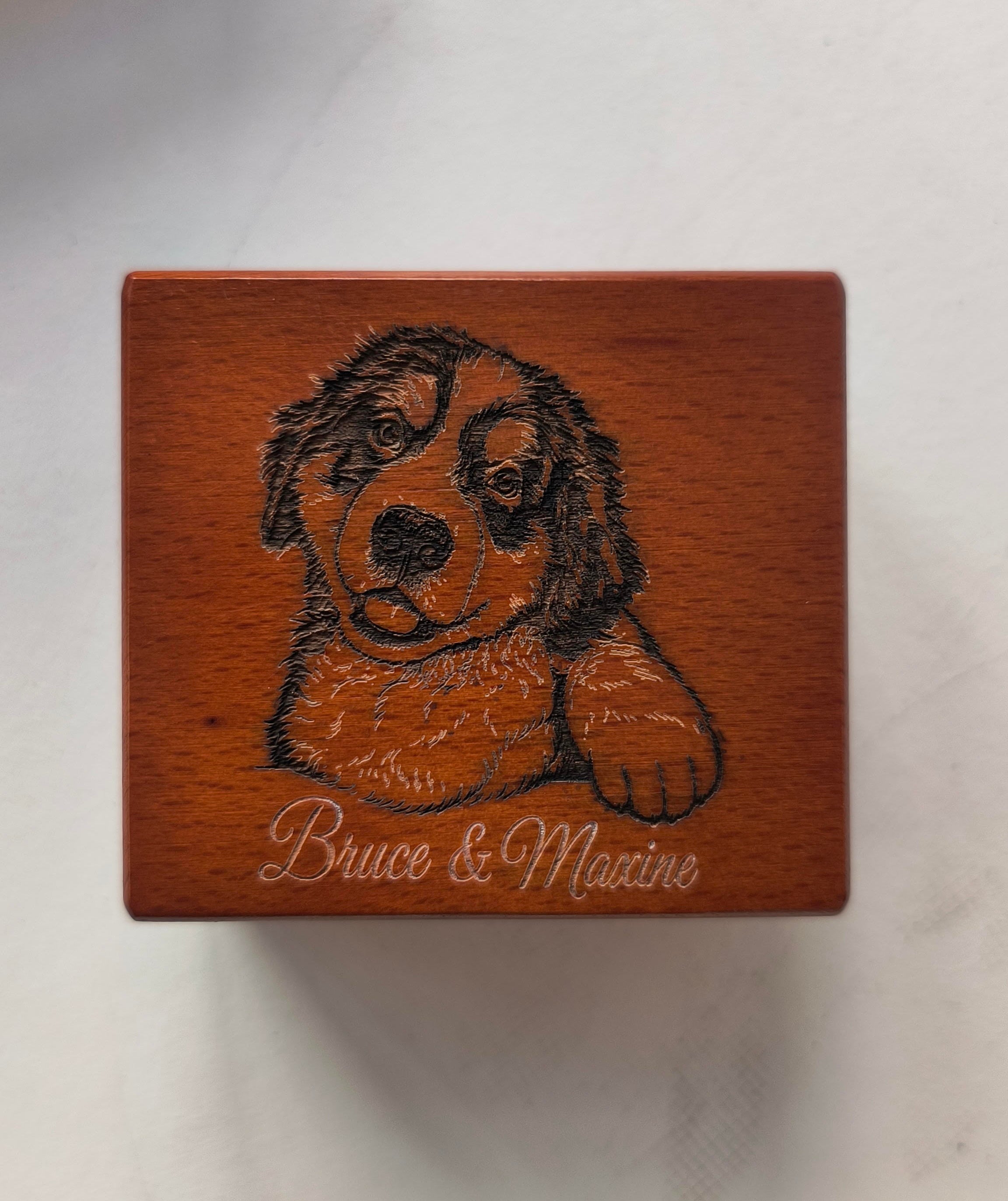 A square wooden music box with a carved portrait of a dog and the engraved text 'Bruce & Maxine' on the front, shown on a light-colored surface. Beautiul custom music box