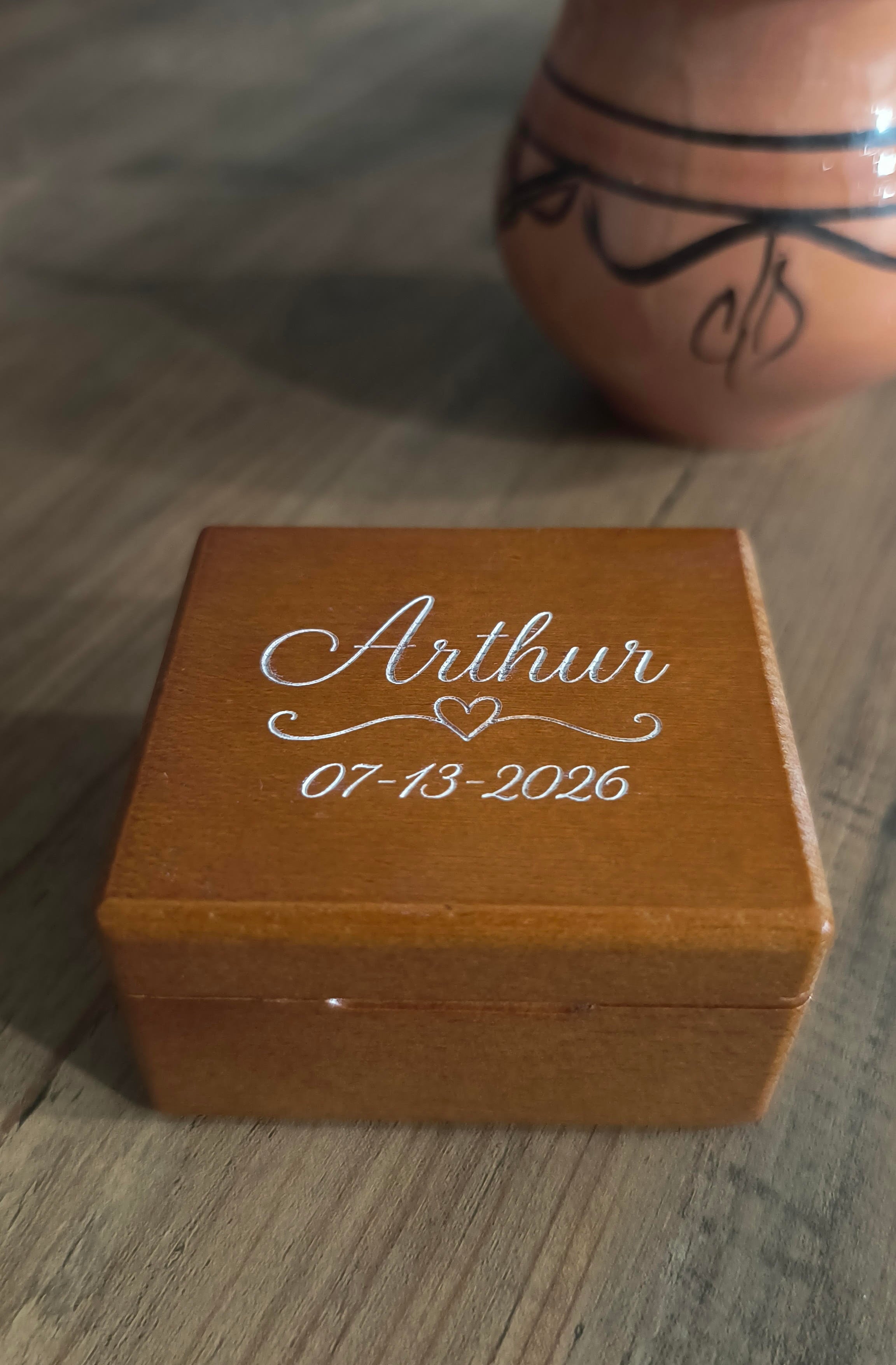 A personalized wooden keepsake box engraved with the name 'Arthur', a heart, and the date '07-13-2026' in white script, placed on a wooden tabletop with a ceramic vase in the blurred background.